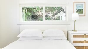 Ein Schlafzimmer mit großem Fenster, weißer Bettwäsche, einer Nachttischlampe und einem gerahmten Bild an der Wand.