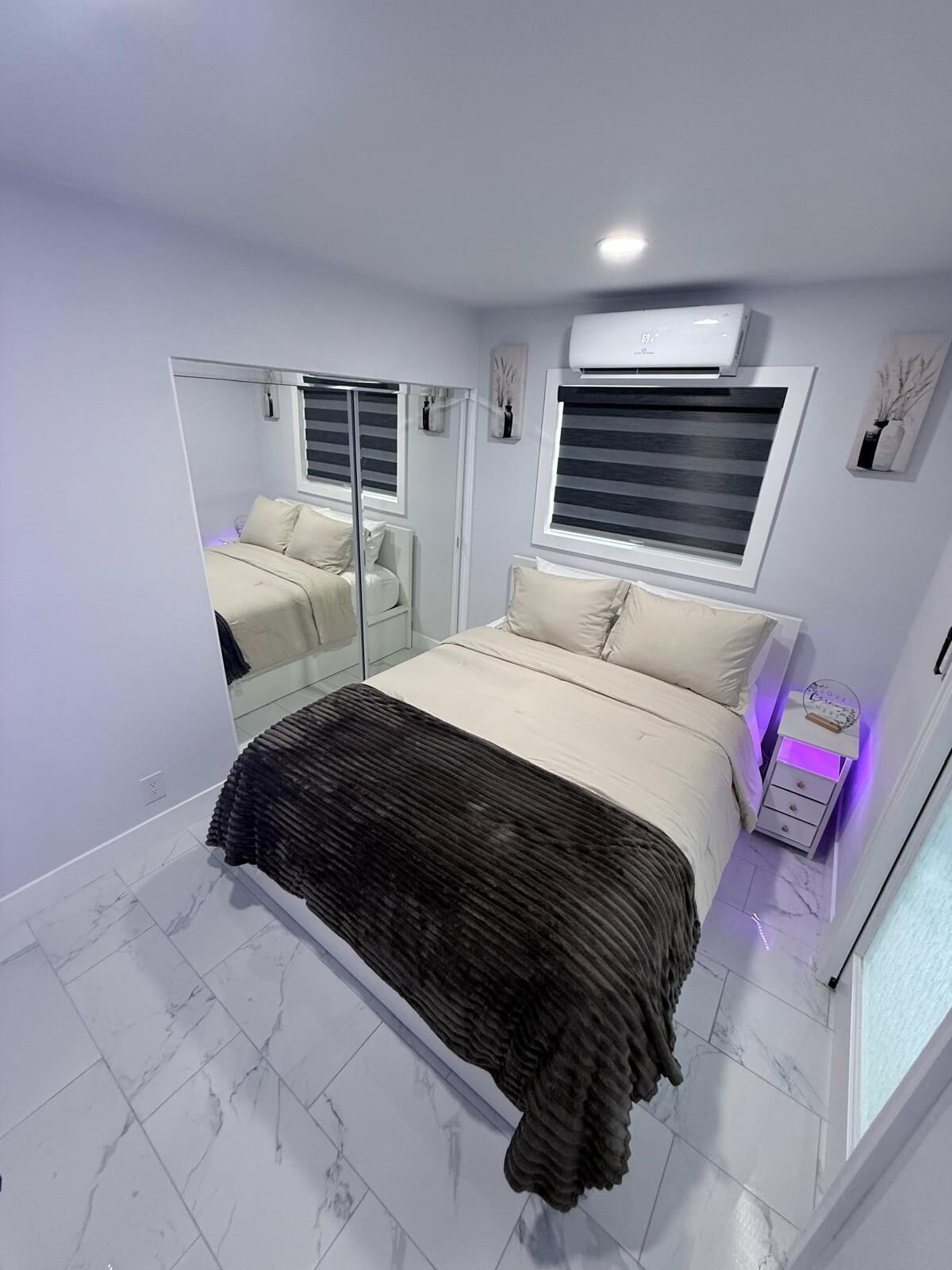 Ein Schlafzimmer mit Bett, Spiegel, Fenster mit Jalousien, Klimaanlage und Nachttisch mit Lampe.