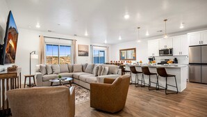 Una sala moderna con un sofá seccional, una mesa de centro y un televisor. Hay una cocina con gabinetes blancos, un microondas y un refrigerador.
