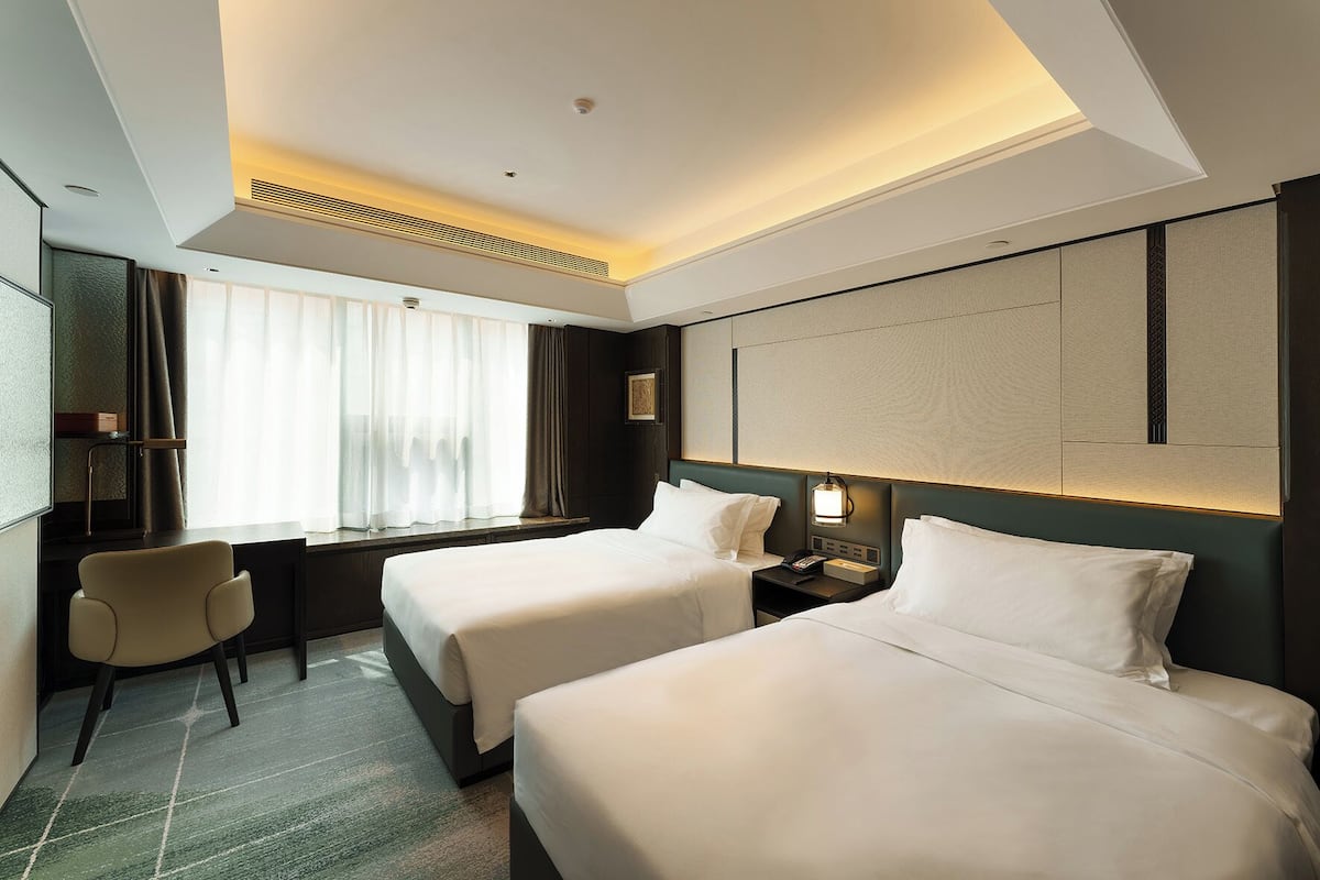Ett hotellrum med två sängar, ett skrivbord, en stol och ett stort fönster med gardiner.