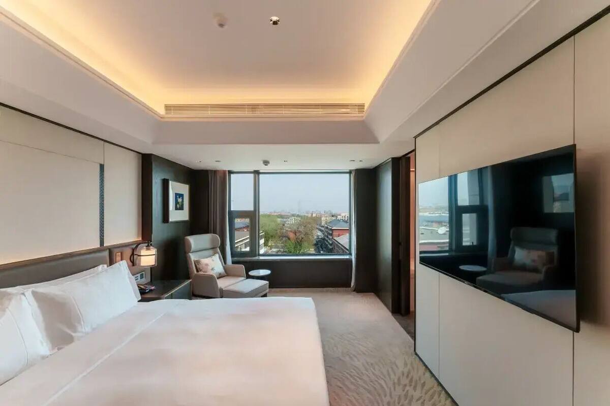 Ett modernt hotellrum med en stor säng, en sittgrupp med en fåtölj och ett litet bord, en platt-TV som är monterad på väggen och ett stort fönster med utsikt över staden.