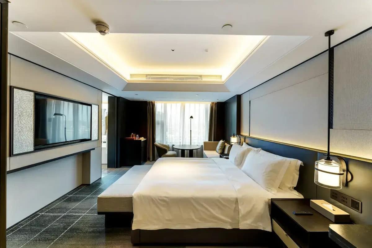 Ett modernt hotellrum med en stor säng, sängbord, ett skrivbord och en stol. Det finns ett stort fönster med gardiner och utsikt över staden.