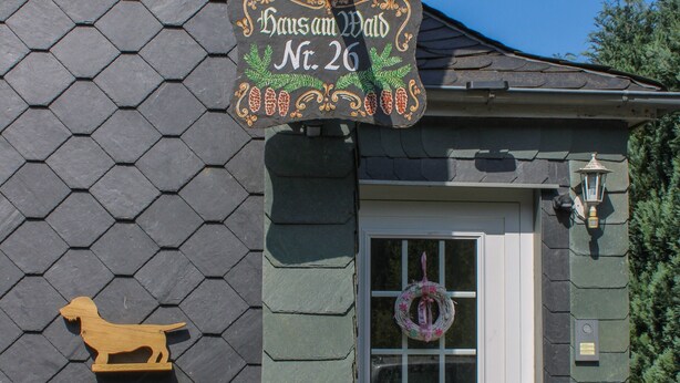 Un bâtiment sur lequel on peut lire l’inscription « Haus am Wald n° 26 », une sculpture de chien en bois, une porte ornée d’une guirlande et une boîte aux lettres.