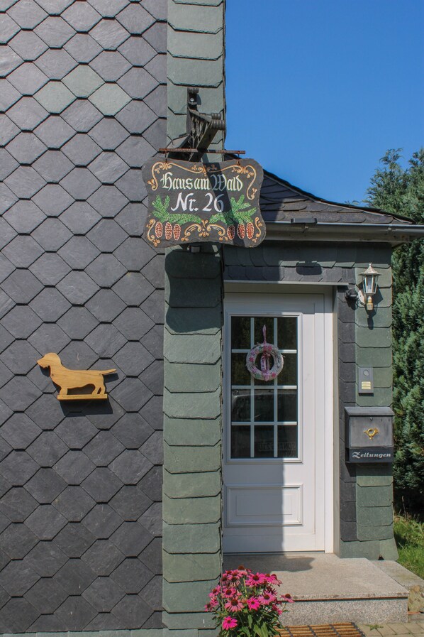 Un bâtiment sur lequel on peut lire l’inscription « Haus am Wald n° 26 », une sculpture de chien en bois, une porte ornée d’une guirlande et une boîte aux lettres.