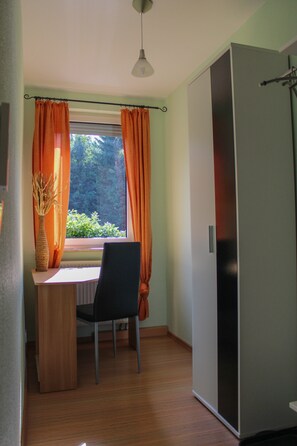 Une pièce avec un plancher en bois, une table avec une chaise, une fenêtre avec des rideaux, un vase contenant des branches séchées et un réfrigérateur.