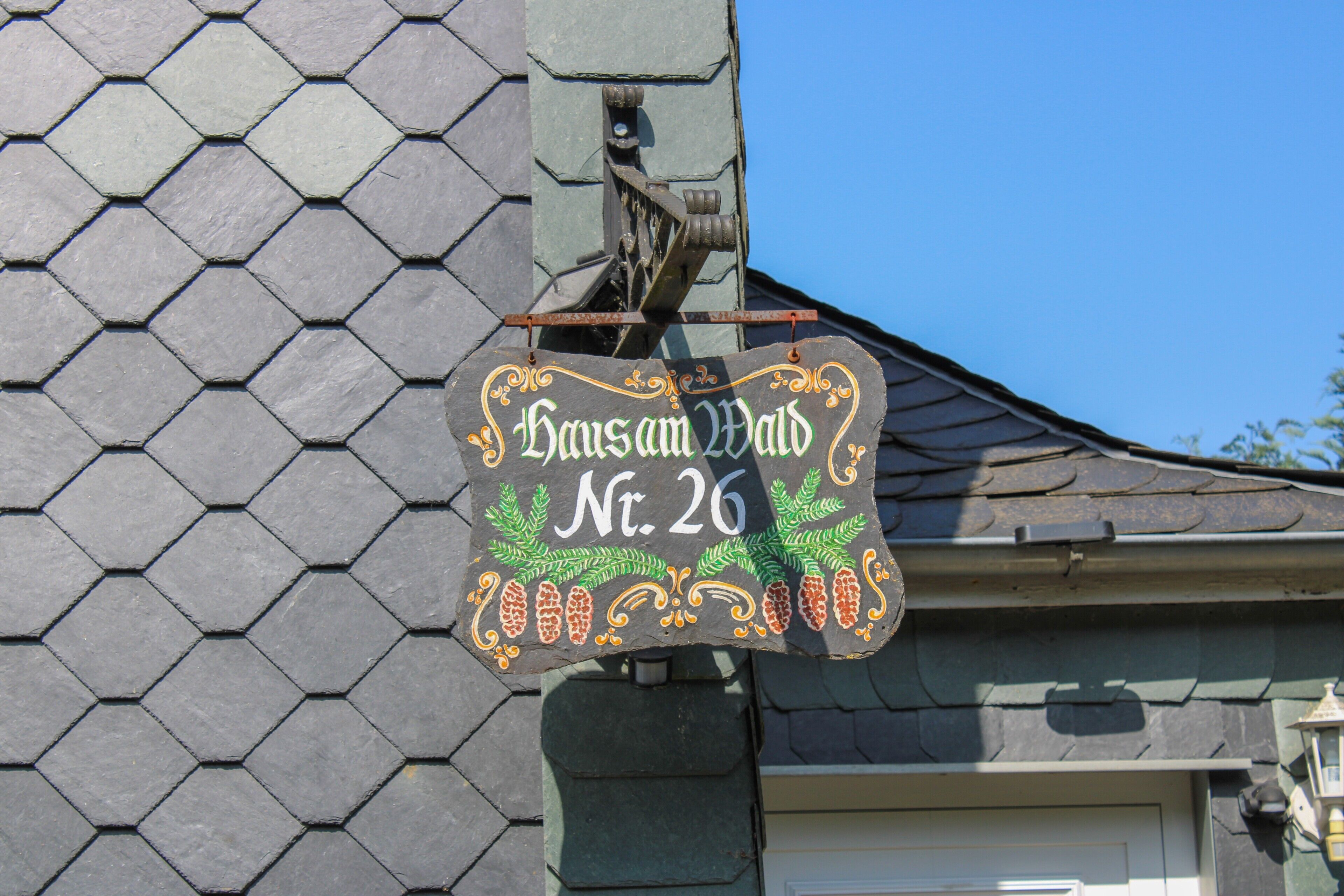 Ein sechseckiges Schild mit der Aufschrift 'Haus am Wald Nr. 26' an der Außenwand eines Gebäudes.