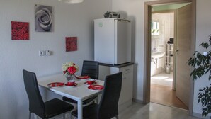 Un coin repas avec une table blanche, des chaises noires et de la vaisselle rouge. On aperçoit un réfrigérateur blanc et un meuble blanc à l’arrière-plan.
