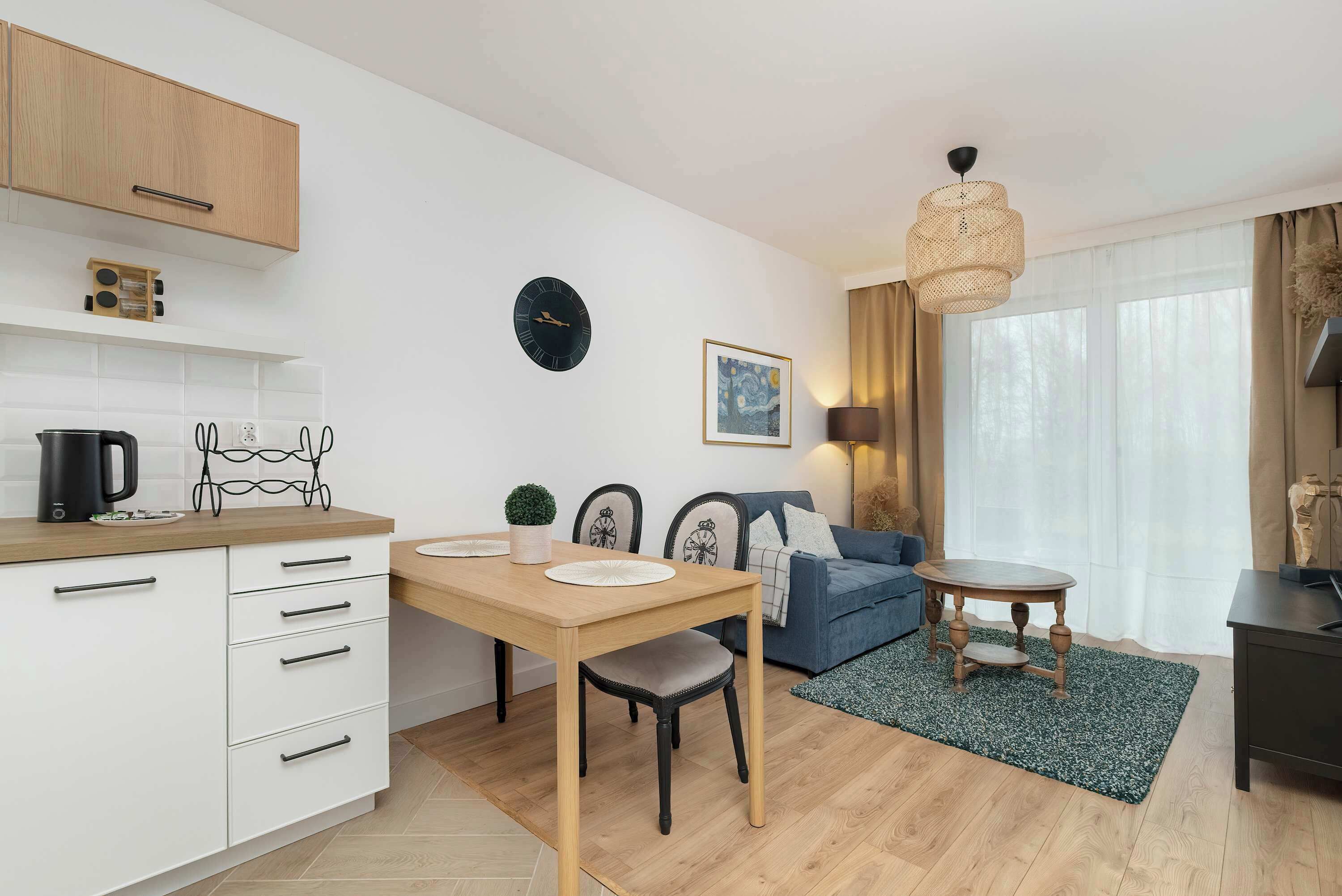 A modern kitchen with white cabinets, a wooden countertop, and a coffee maker. There is a dining table with chairs, a blue sofa, and a small wooden table with a lamp. A clock and a framed picture are on the wall.