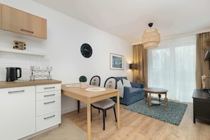 Eine moderne Küche mit weißen Schränken, einer Holzarbeitsplatte und einer Kaffeemaschine. Es gibt einen Esstisch mit Stühlen, ein blaues Sofa und einen kleinen Holztisch mit einer Lampe. An der Wand hängen eine Uhr und ein gerahmtes Bild.