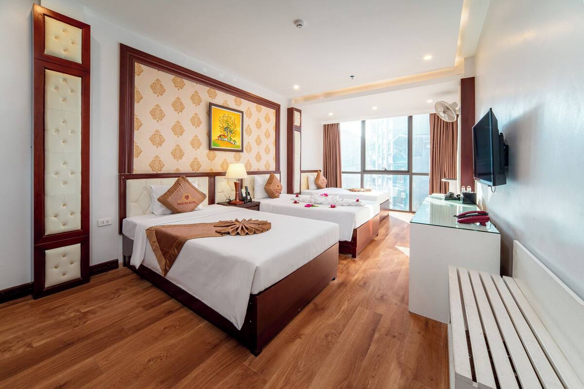 A hotel room with three beds, a TV, a desk, and a large window.