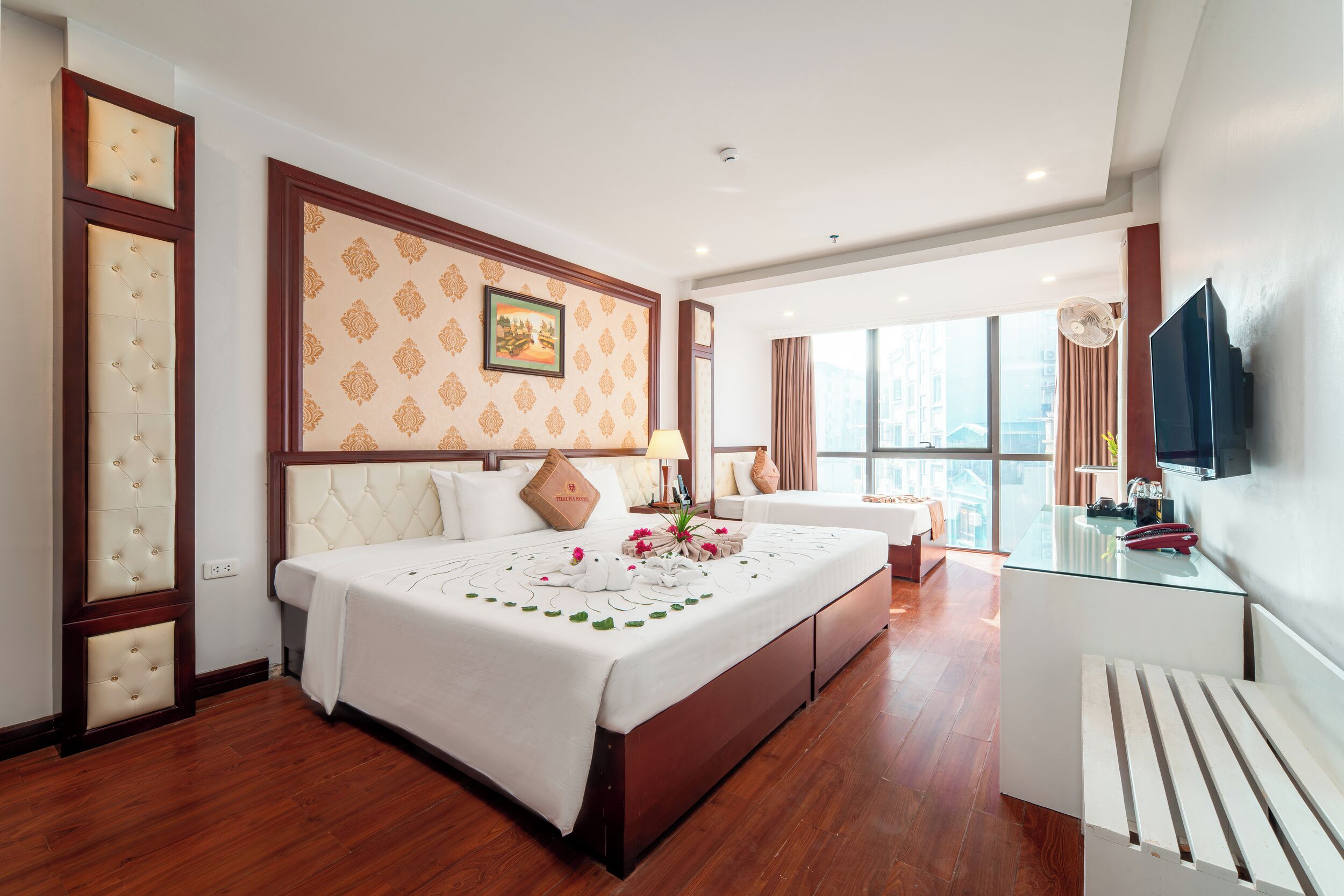 A hotel room with a large bed, a glass table, a TV, and a view of the city.