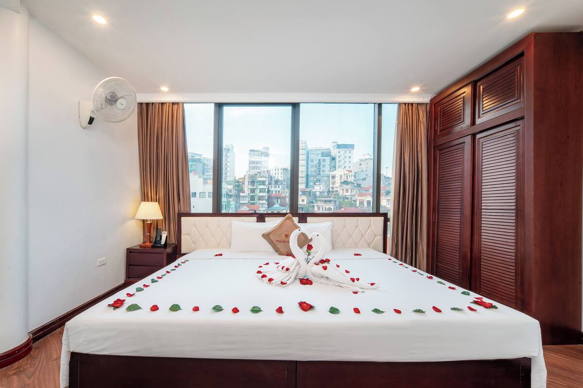A hotel room with a large bed, a nightstand, a wall-mounted fan, and a view of the cityscape through large windows.