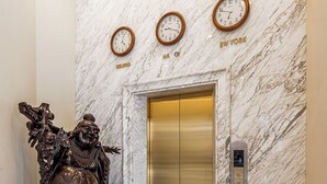 A marble wall with clocks displaying Beijing, Ha Noi, and New York times, an elevator, and a statue. - Thai Ha Hotel (Bai Chay)