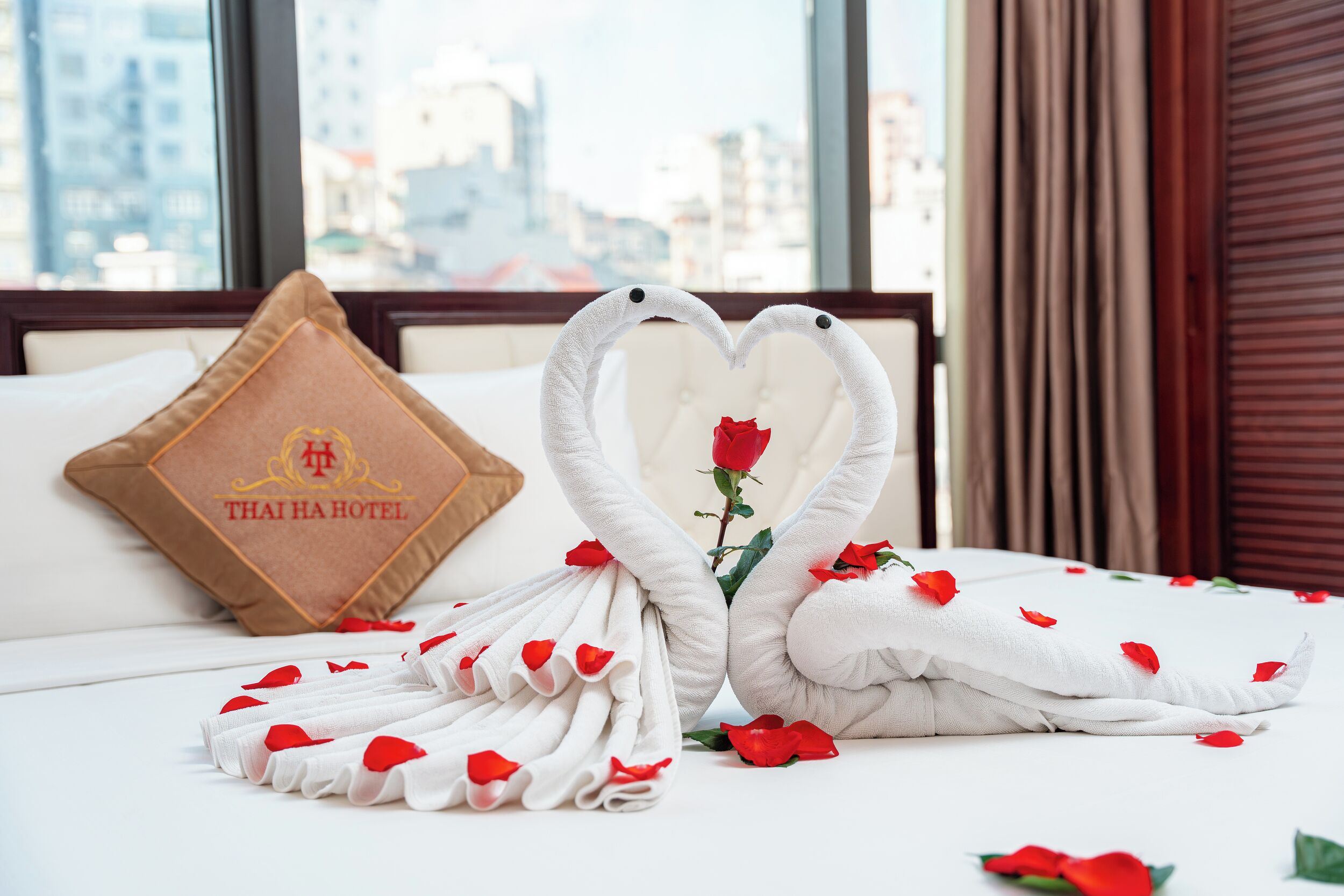 Ein Bett mit einer Herz-Anordnung aus Handtüchern, einer Rose und einem Kopfkissen mit dem Aufdruck "THAI HA HOTEL".
