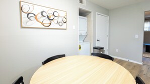 Une table ronde en bois, accompagnée de quatre chaises, une œuvre d’art abstraite accrochée au mur, une buanderie équipée d’un lave-linge et d’un sèche-linge, et une porte donnant sur une autre pièce.