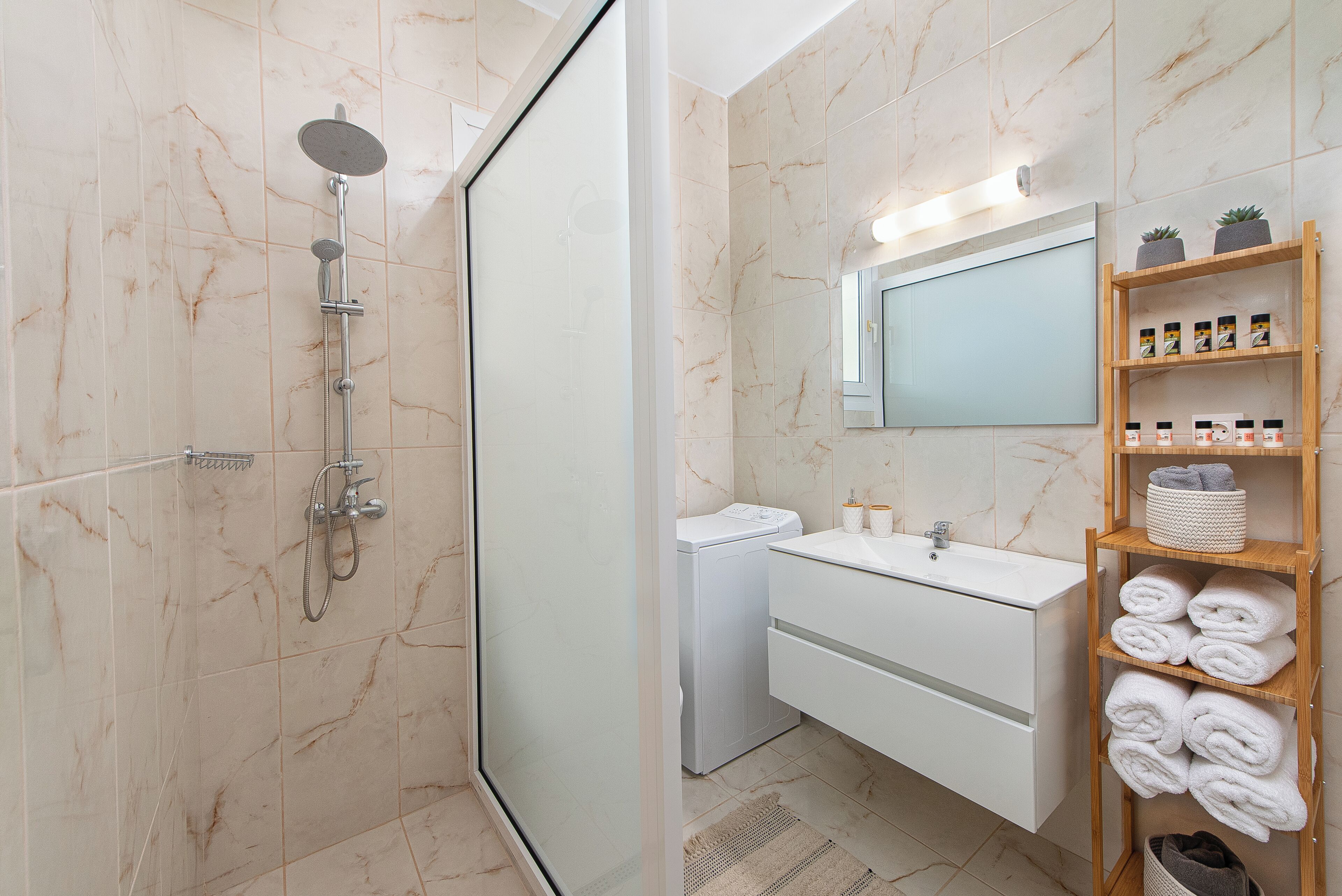Une salle de bain moderne avec une douche à parois de verre, un lavabo blanc, un miroir et une étagère en bois où sont posées des serviettes.