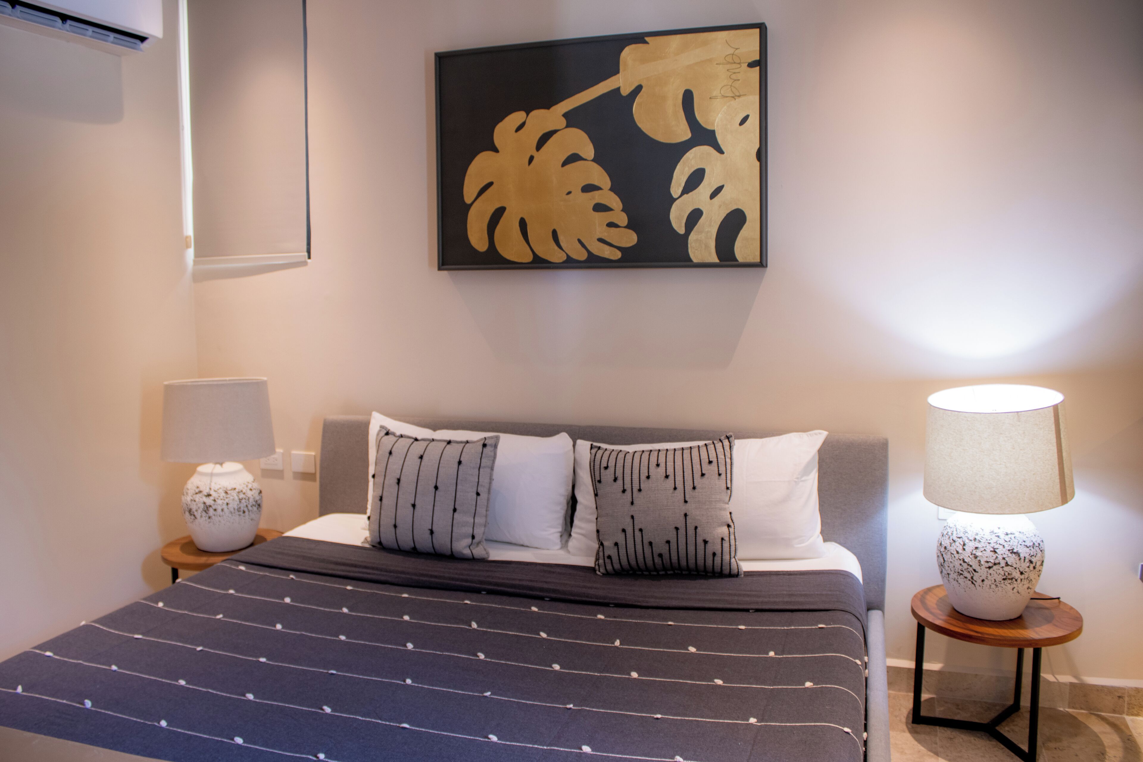 Un dormitorio ordenado con una cama, almohadas, una mesita de noche con una lámpara y una obra de arte enmarcada en la pared.
