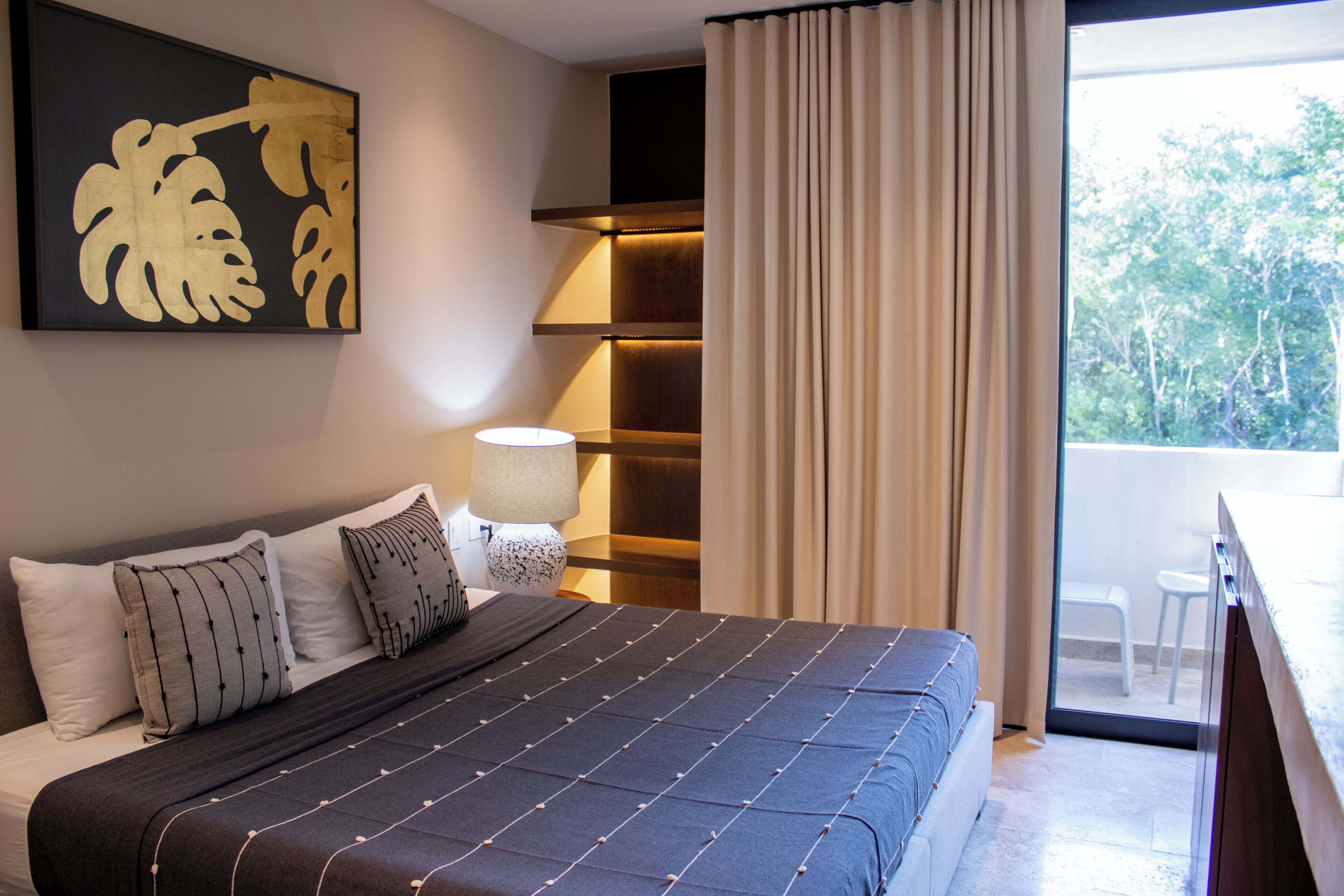 A modern hotel room with a large bed, a bedside table with a lamp, a view of the outdoors, and a large abstract painting on the wall.