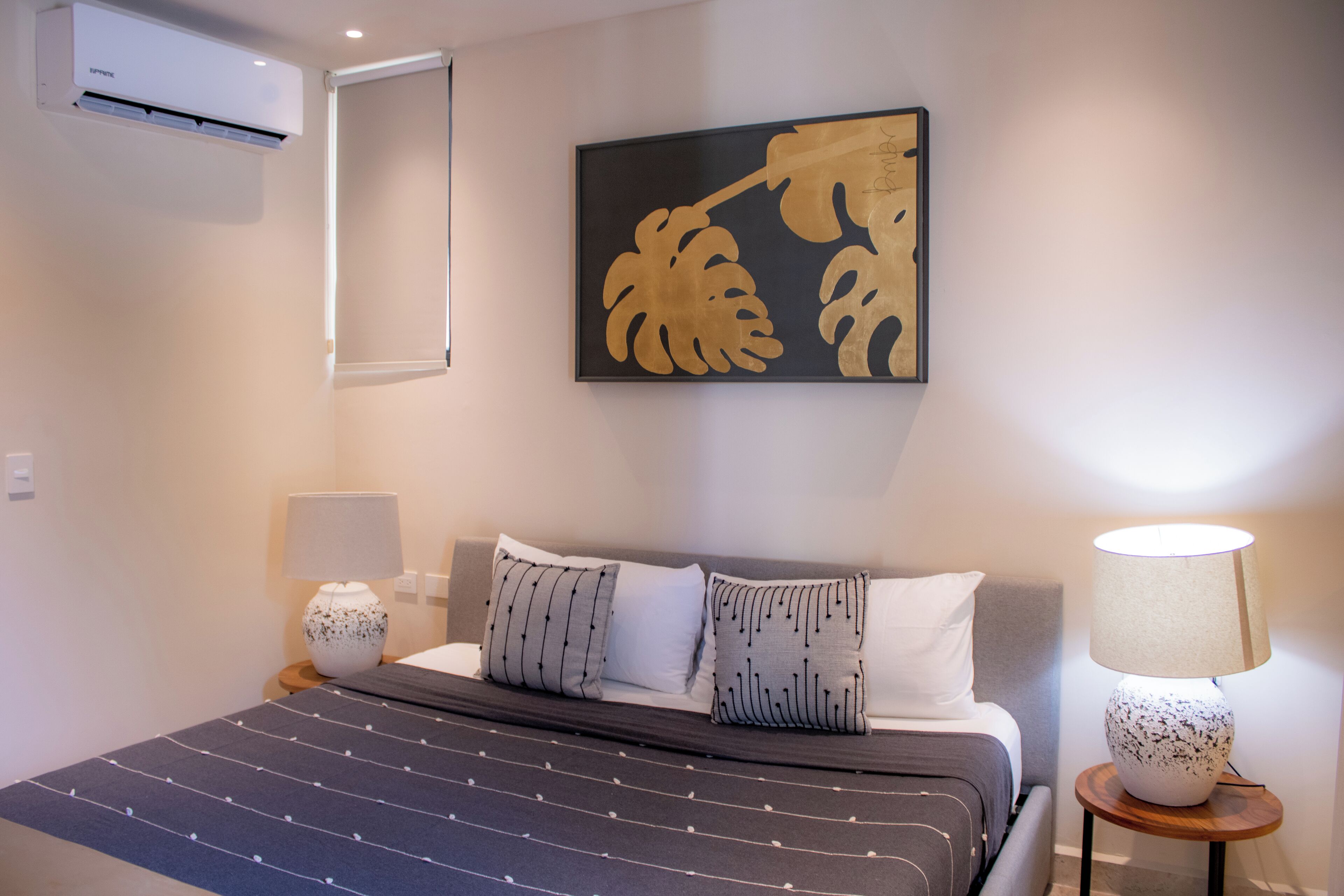 Un dormitorio ordenado con cama, almohadas, una mesita de noche, lámpara y aire acondicionado de pared.