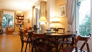 Ein Essbereich mit einem runden Tisch, Stühlen und einer Lampe. Ein Bücherregal und ein Fenster mit Vorhängen sind ebenfalls vorhanden.