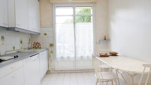 Eine Küche mit weißen Schränken, einer Arbeitsplatte, einem Spülbecken, einem Fenster mit weißem Vorhang und einem Esstisch mit Stühlen.