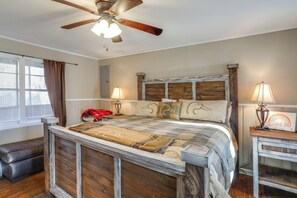Un dormitorio con estructura de cama de madera, ventilador de techo, mesita de noche con lámpara y un banco.