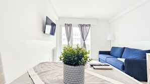 A modern living room with a blue sofa, a grey textured coffee table, a potted plant, a flat-screen TV mounted on the wall, and a window with grey curtains. - New Big Flat 8 Pers AC WiFi (Nice)