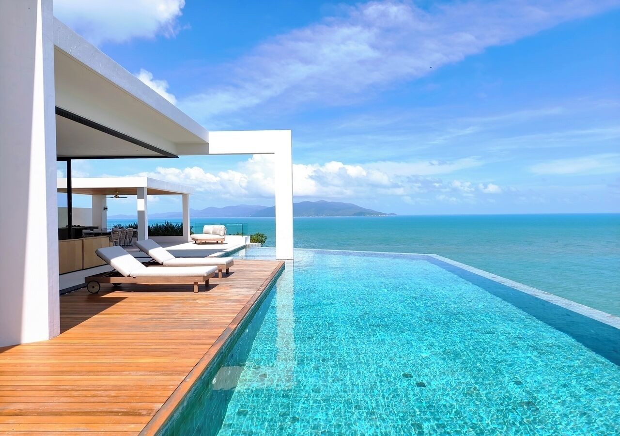 Piscina infinita moderna con terraza de madera y sillones de descanso, con vistas al mar azul y montañas a lo lejos.