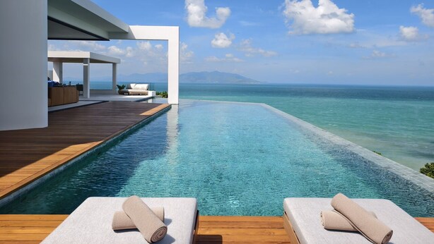 Una piscina infinita moderna con cubierta de madera y sillones de descanso, con vista a un mar azul y montañas distantes.