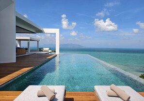 Une piscine à débordement moderne, dotée d’une terrasse en bois et de chaises longues, offrant une vue imprenable sur une mer d’un bleu profond et sur des montagnes lointaines.