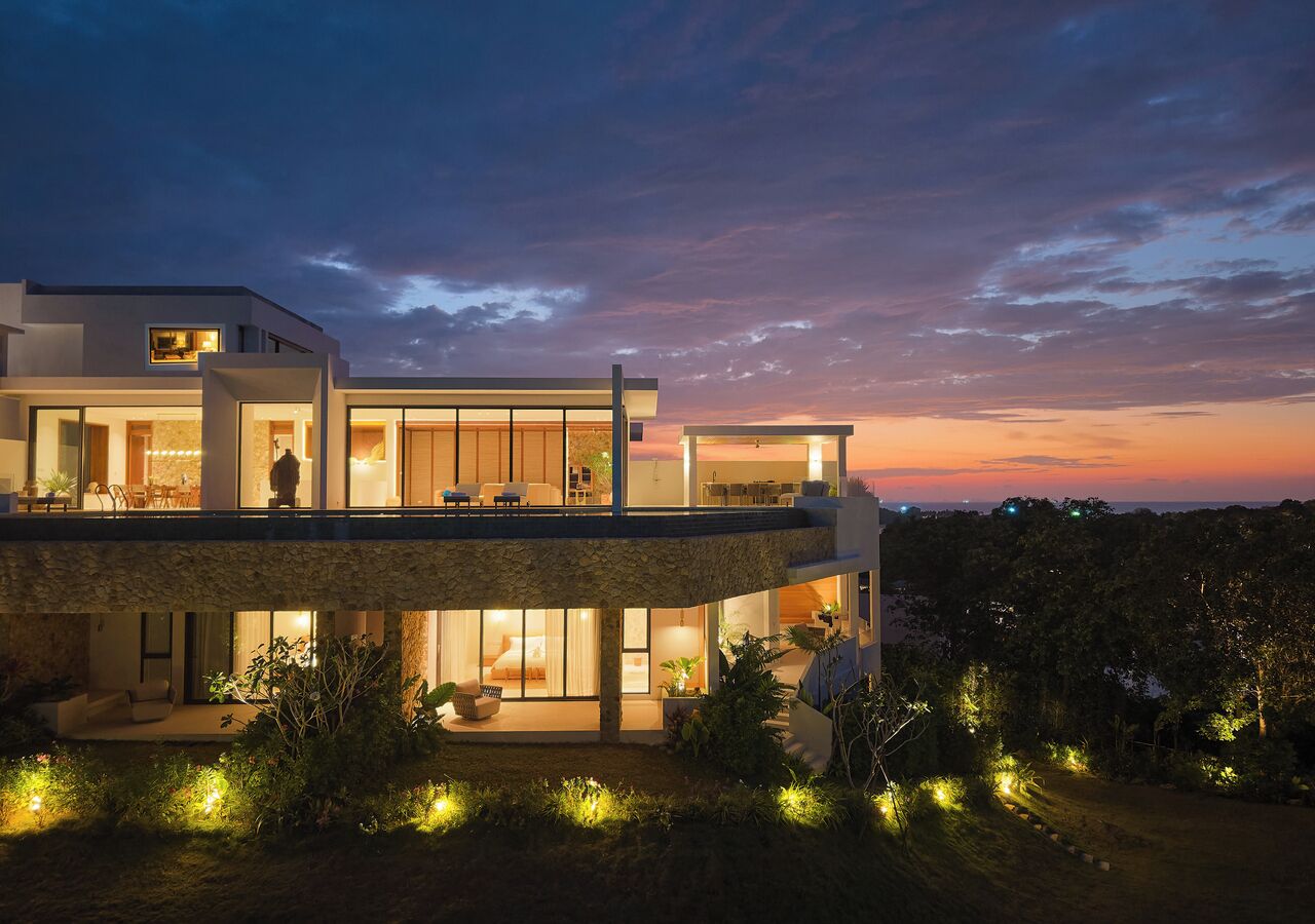A modern two-story house with a balcony, large windows, and outdoor lighting.