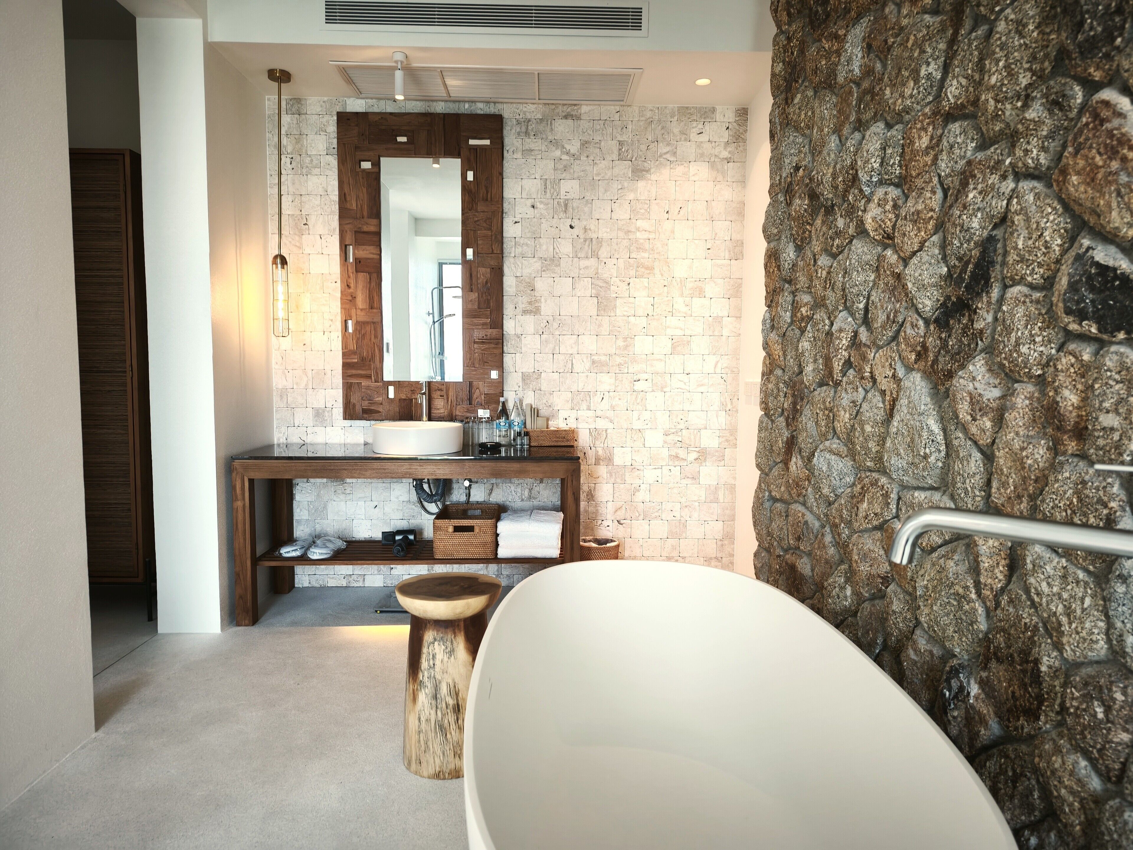 A modern bathroom with a stone wall, a wooden vanity, a large mirror, and a freestanding bathtub.