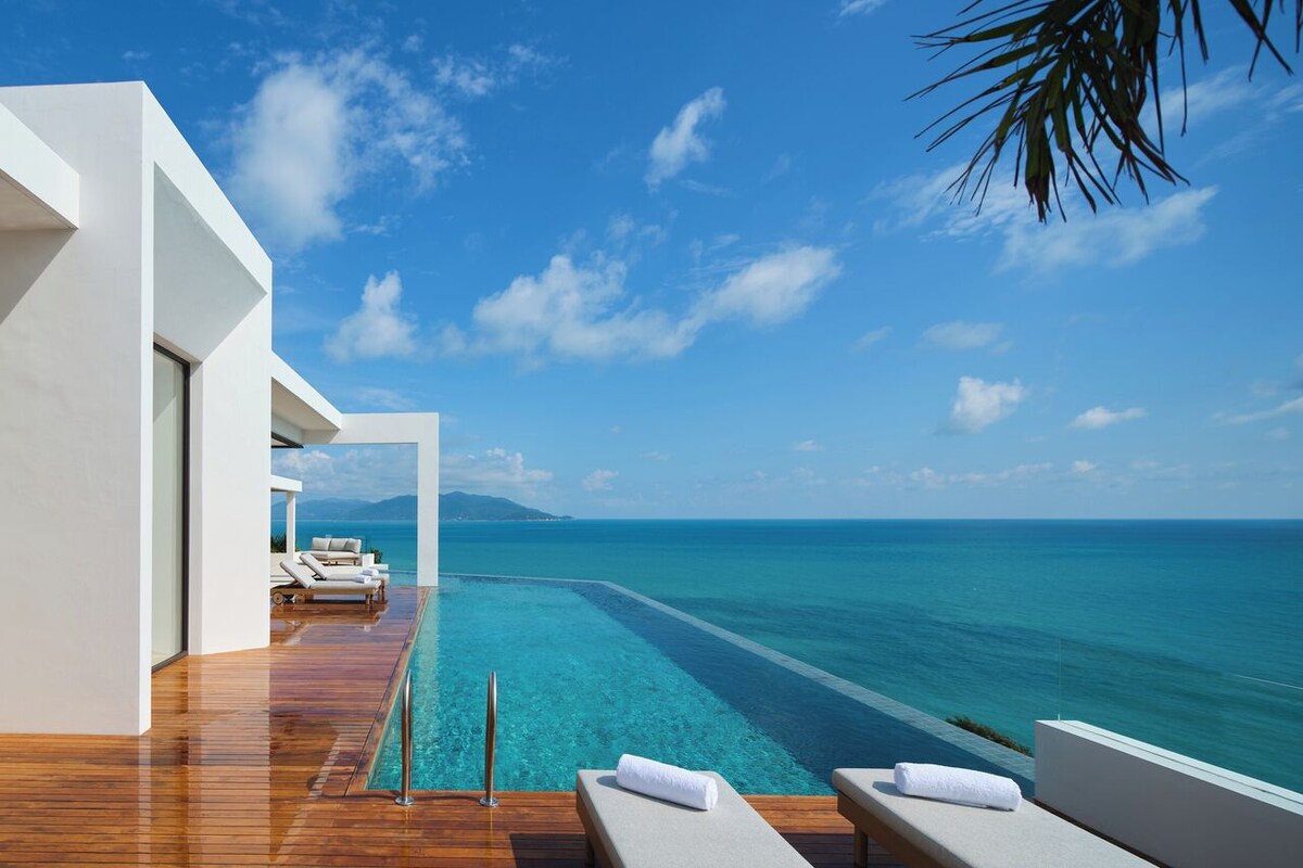 An infinity pool with a clear view of the ocean, a wooden deck, and a white building structure.