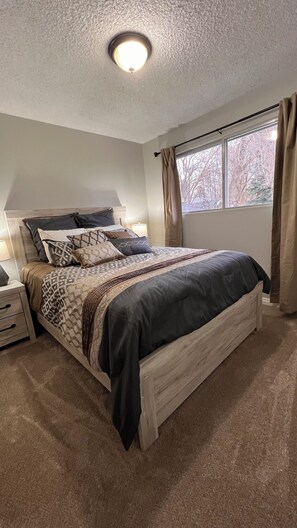 Un dormitorio con cama, mesitas de noche, ventana con cortinas y una lámpara en el techo.