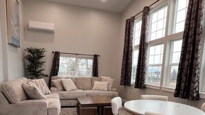 Ein Wohnzimmer mit Ecksofa, Couchtisch und Esstisch mit Stühlen. Ein Fenster mit Vorhängen, eine Klimaanlage an der Wand und eine Pflanze sind ebenfalls vorhanden.