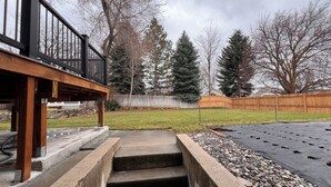 Ein Garten mit einer Holzterrasse, einer Betonstiege, einem Rasen, einem Holzzaun und Bäumen.