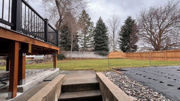 Un jardin avec une terrasse en bois, un escalier en béton, une pelouse, une clôture en bois et des arbres.