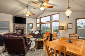 A spacious living room with a fireplace, leather sofas, a dining table, and large windows offering a view of the outdoors. - Direct Riverfront Home w/ Private Dock, Game Room, Deck, Amazing Views, Just 4 Miles to Beach (Nehalem)