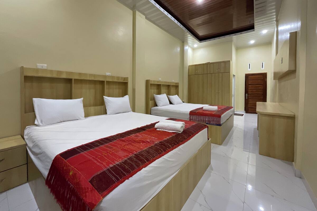 A hotel room with two beds, wooden headboards, a red and white bedspread, a wooden wardrobe, and a tiled floor.
