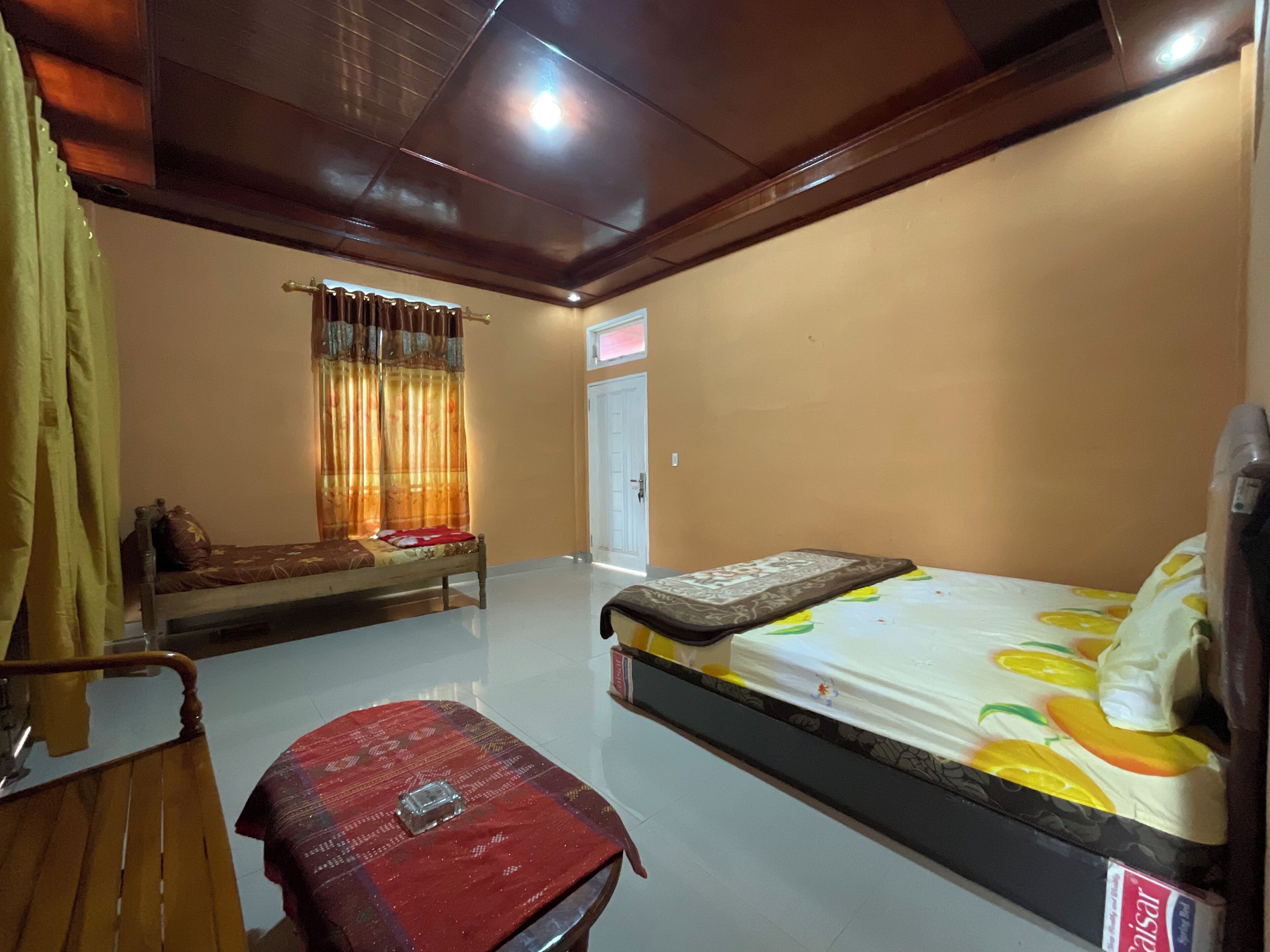 Quarto com cama, cadeira de madeira, mesa pequena e janela com cortinas.