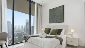 A modern bedroom with a large bed, a nightstand, a chair, and a city view through floor-to-ceiling windows. - Binghatti Royale Residences - JVC (Dubai)