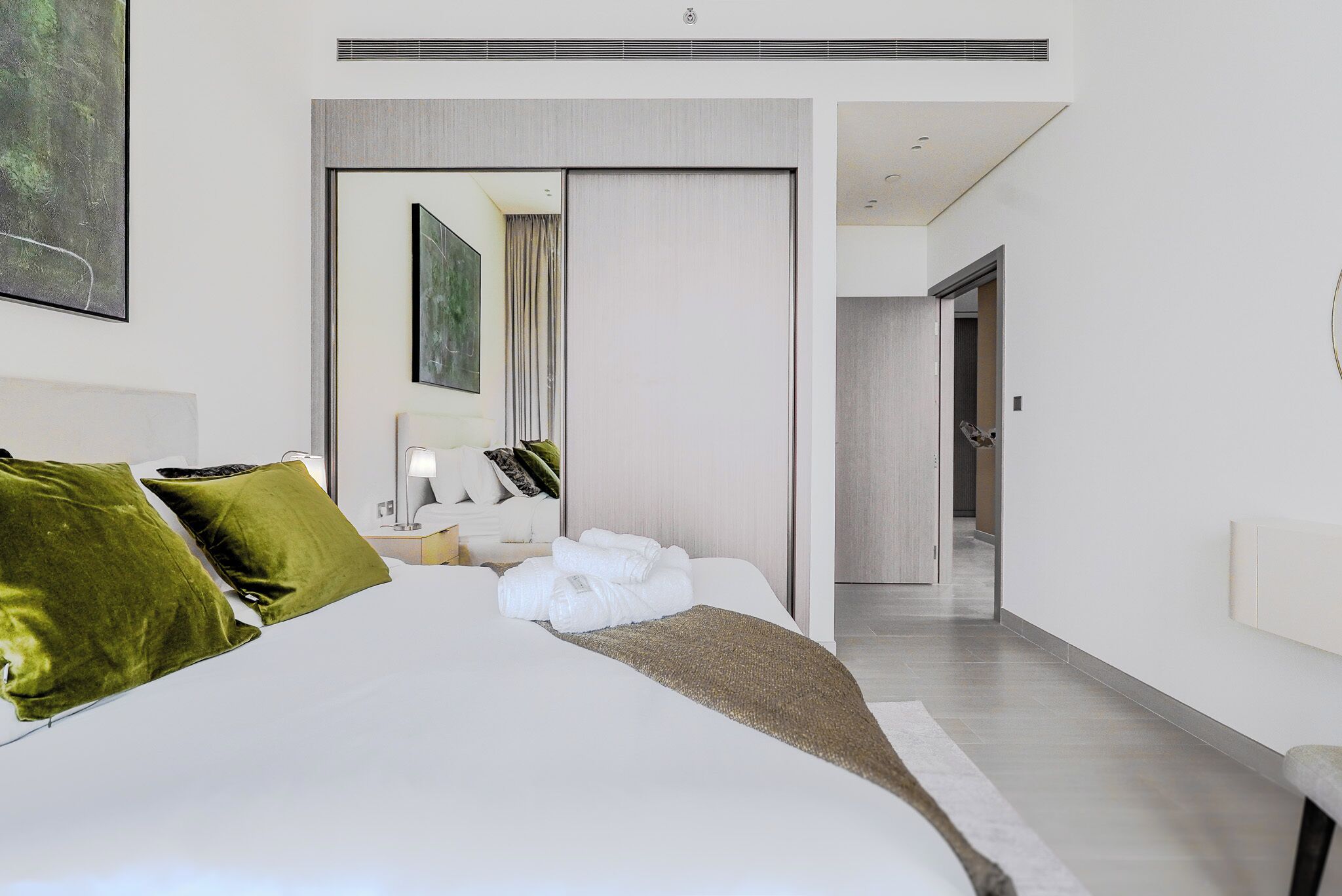 Una camera d'albergo moderna con un letto grande, cuscini verdi, un comodino con una lampada e vista sul corridoio.