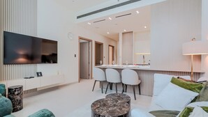 A modern living room with a flat-screen TV, a dining area with chairs, a kitchen with a bar, and a sofa with cushions. - Binghatti Royale Residences - JVC (Dubai)
