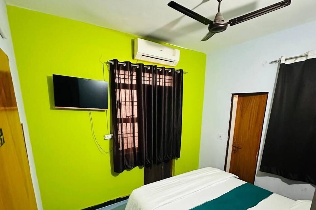 A room with a bed, a ceiling fan, a TV mounted on the wall, and a window with curtains.