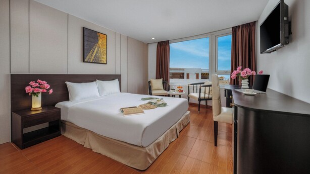 Habitación de hotel con una cama, una mesita de noche con flores, un escritorio con una computadora portátil, un televisor y vistas al cielo.