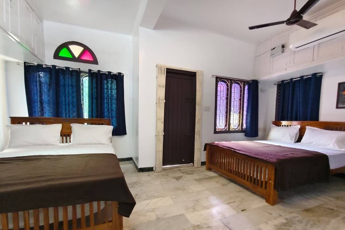 Une chambre d’hôtel avec deux lits, une tête de lit en bois, un ventilateur de plafond, une porte avec une fenêtre et un climatiseur fixé au mur.
