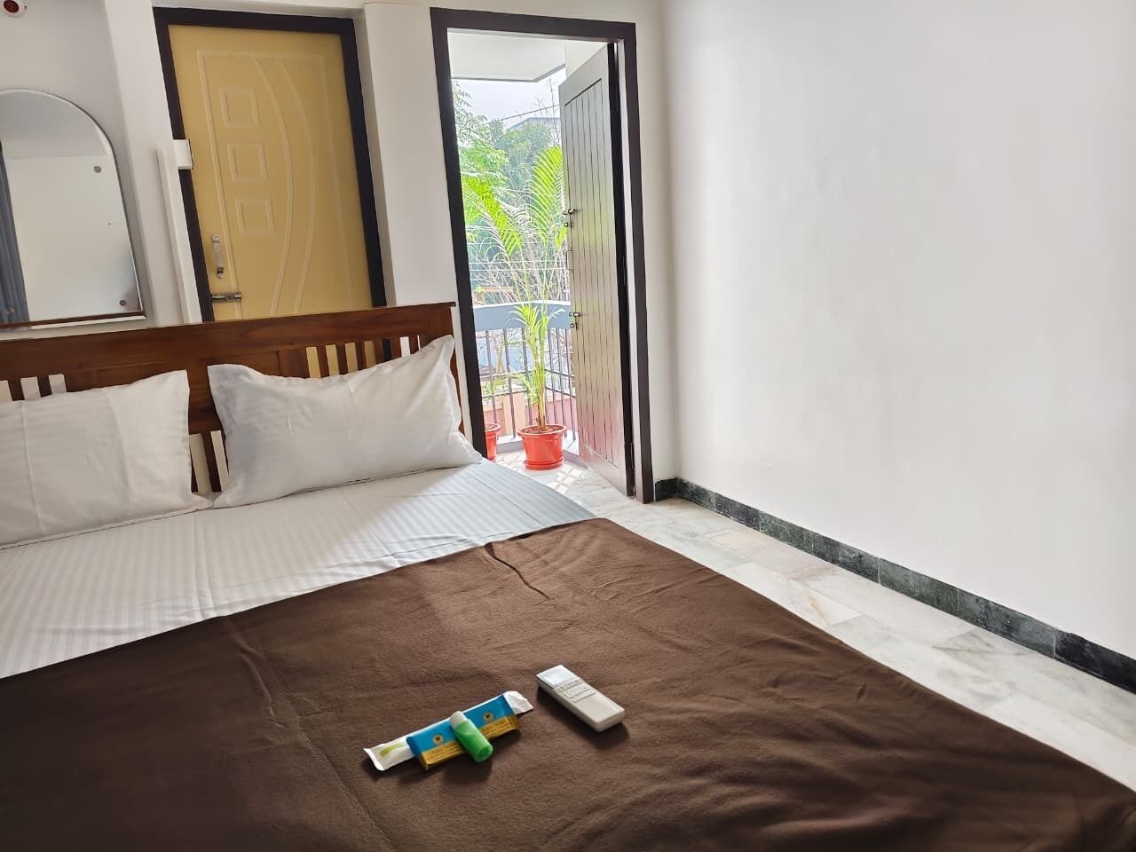 A hotel room with a bed, a wooden headboard, a brown bedspread, and a window with a view of greenery outside.