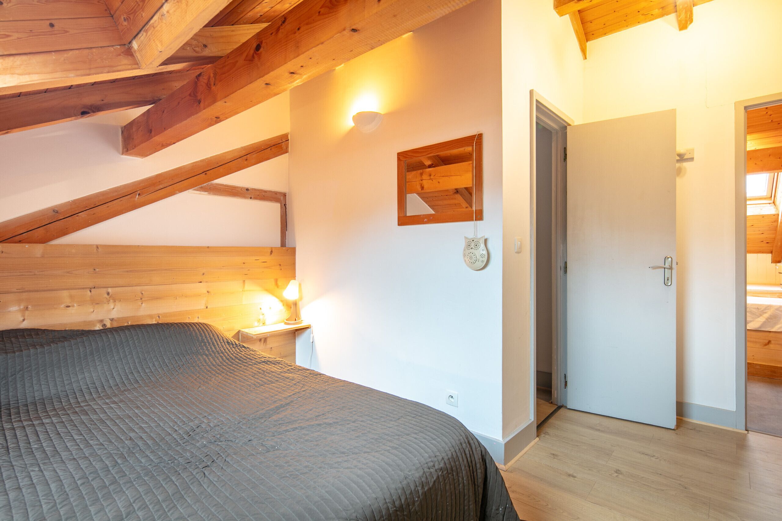Ein Schlafzimmer mit Holzboden, einem Bett, einem Nachttisch mit Lampe, einem an der Wand befestigten Spiegel und einer Tür zu einem anderen Raum.