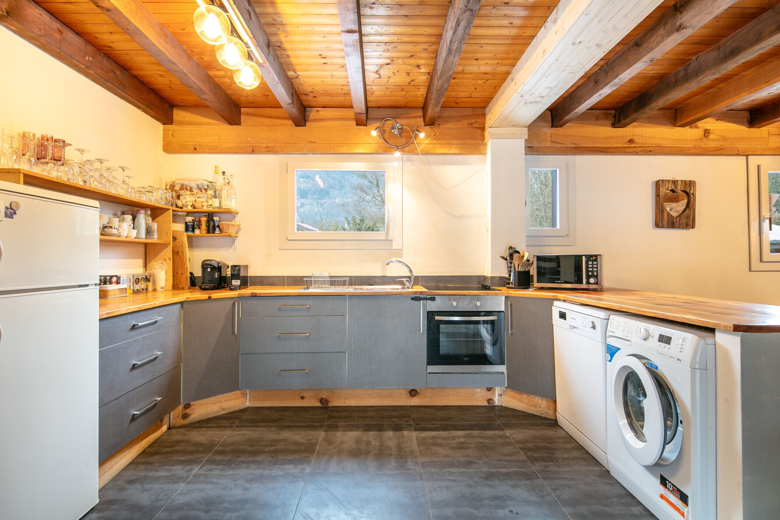 Une cuisine compacte avec des poutres en bois, un four encastré, un lave-linge et une fenêtre laissant entrer la lumière naturelle.