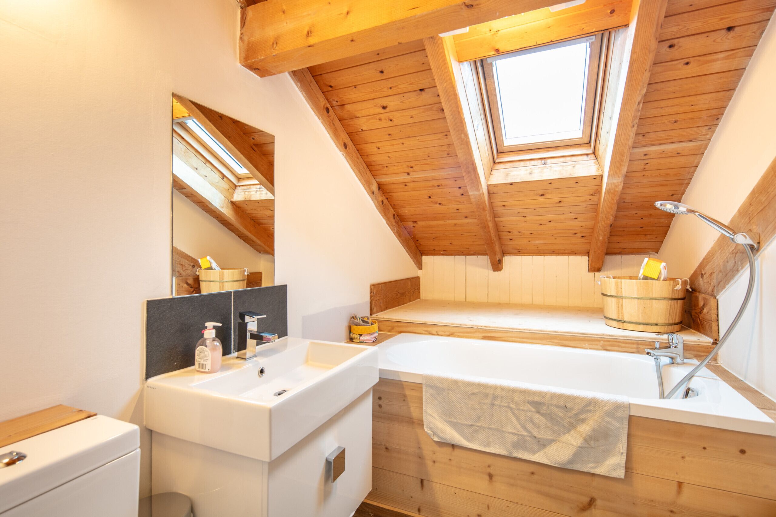 A modern bathroom with a skylight, wooden beams, a bathtub, and a vanity with a mirror.